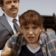 Millie Bobby Brown's Eleven, looking quite a bit older and wearing a flannel shirt, is being held back by the arms by two men in grey suits.